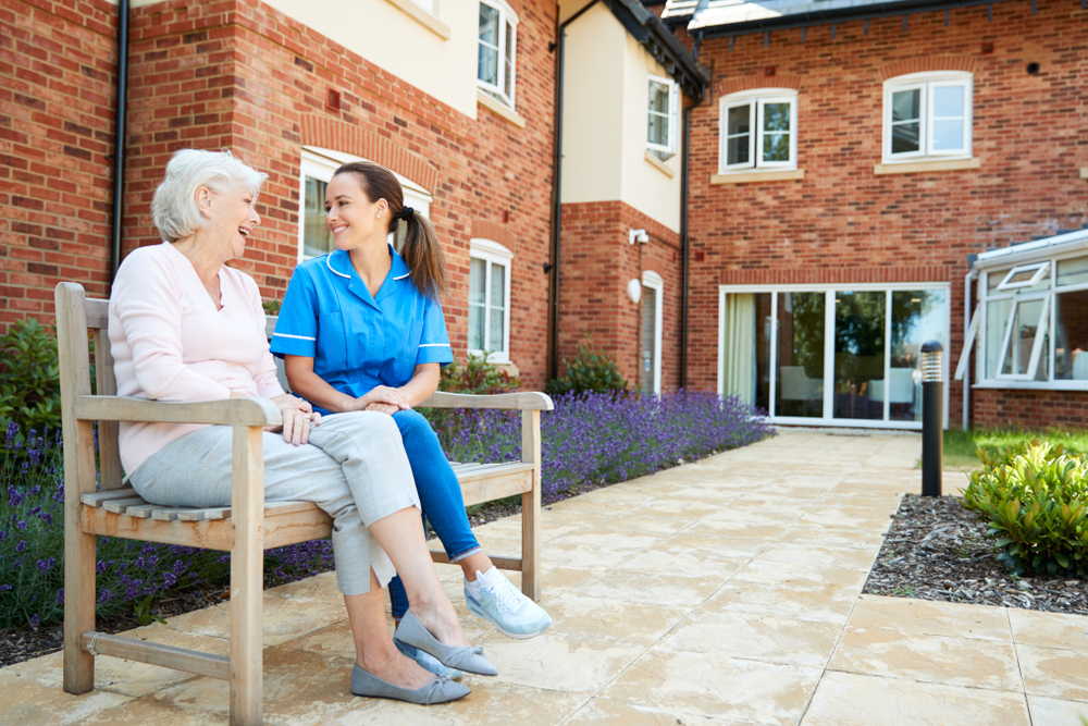 Senior Woman Sitting On Bench And Talking With Nurse In Retirement Home